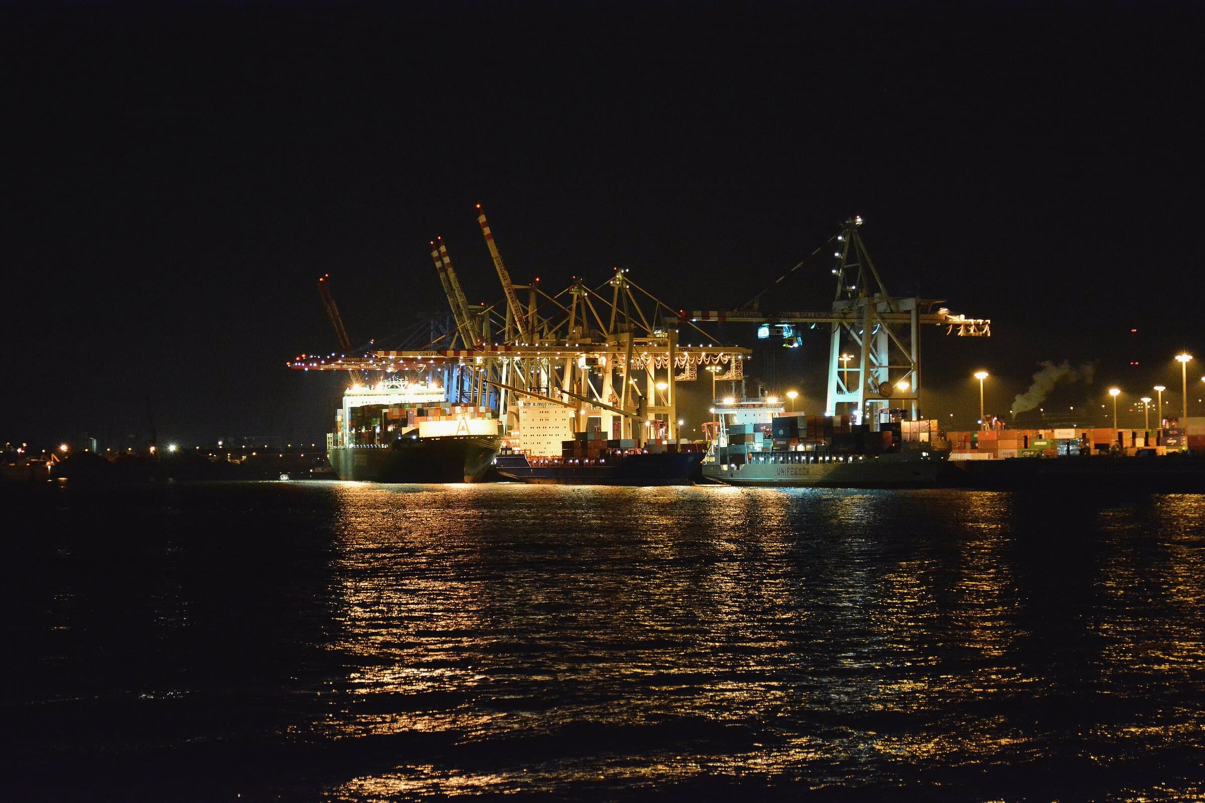 A container terminal at night, gantry cranes lit above a moored vessel, lights reflected in the water.